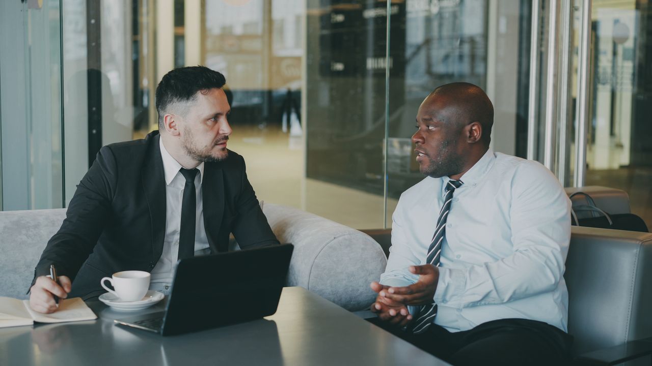 Mastering the First Impression: Your intriguing post title goes here Two businessmen discussing at a cafe with a laptop and coffee.