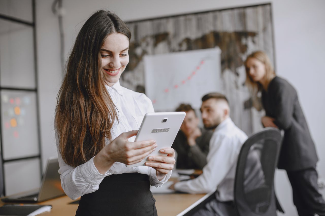 Young woman in a professional office setting browsing a tablet with colleagues in the background.
